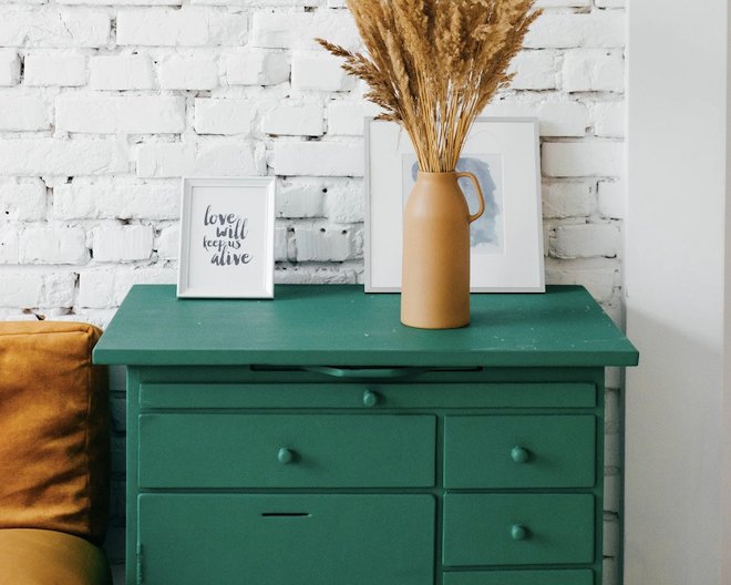 A vase, a frame with the quote 'love will keep us alive', and a framed sketch resting on a teal desk with drawers in front of a white brick wall