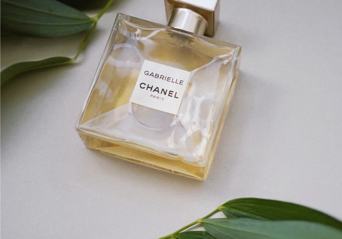 A bottle of Gabrielle Essence Eau De Parfum lying flat on a white surface with leaves around it